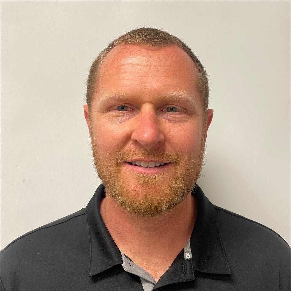 A man with short light brown hair and a reddish beard is smiling slightly. He is wearing a black collared shirt, representing Paine Excavating, and is posed against a plain white background, highlighting his expertise in excavation services.
