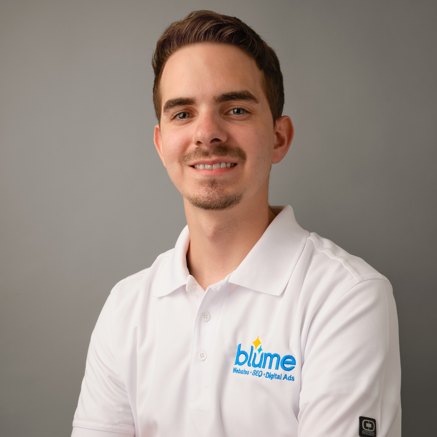 A young man with short brown hair and a trimmed beard is wearing a white polo shirt with the "blume Website • SEO • Digital Ads" logo. He is smiling and posing in front of a plain gray background.