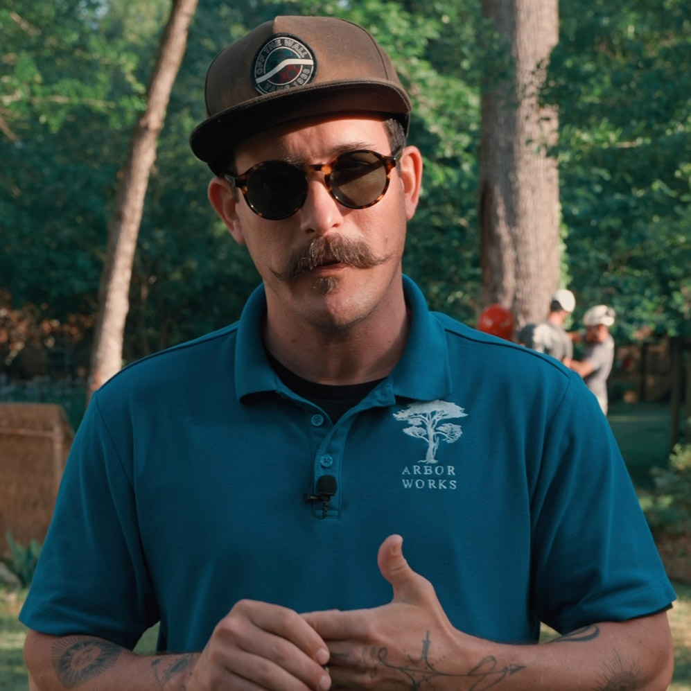 A man wearing sunglasses, a cap, and a blue "Arbor Works" polo shirt stands outdoors. There are trees and people in the background. He has a mustache and is looking at the camera with his hands together.