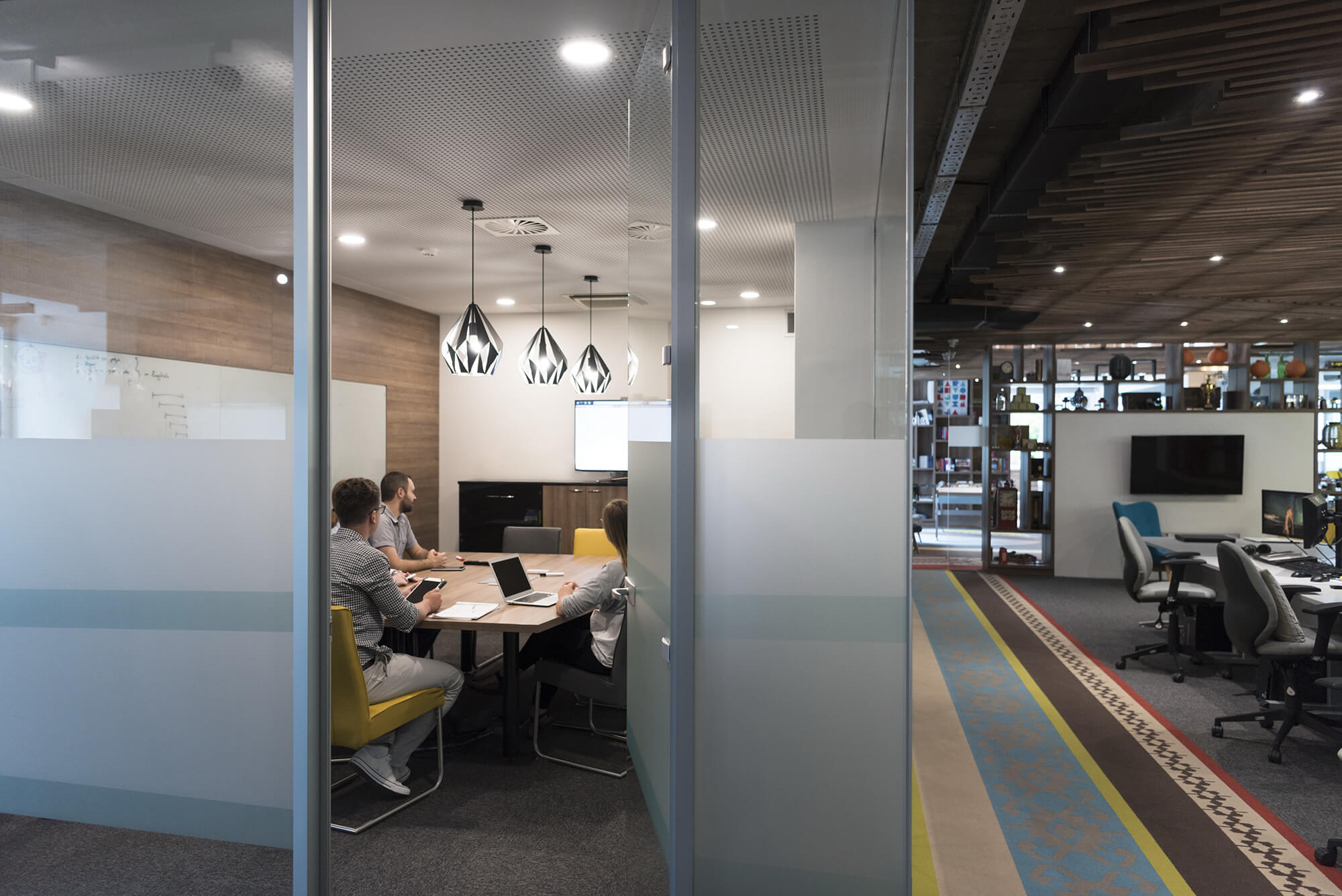 Three people sit at a conference table in a glass-walled meeting room, looking at a screen with a presentation. The adjacent open office area has desks, chairs, and computers.