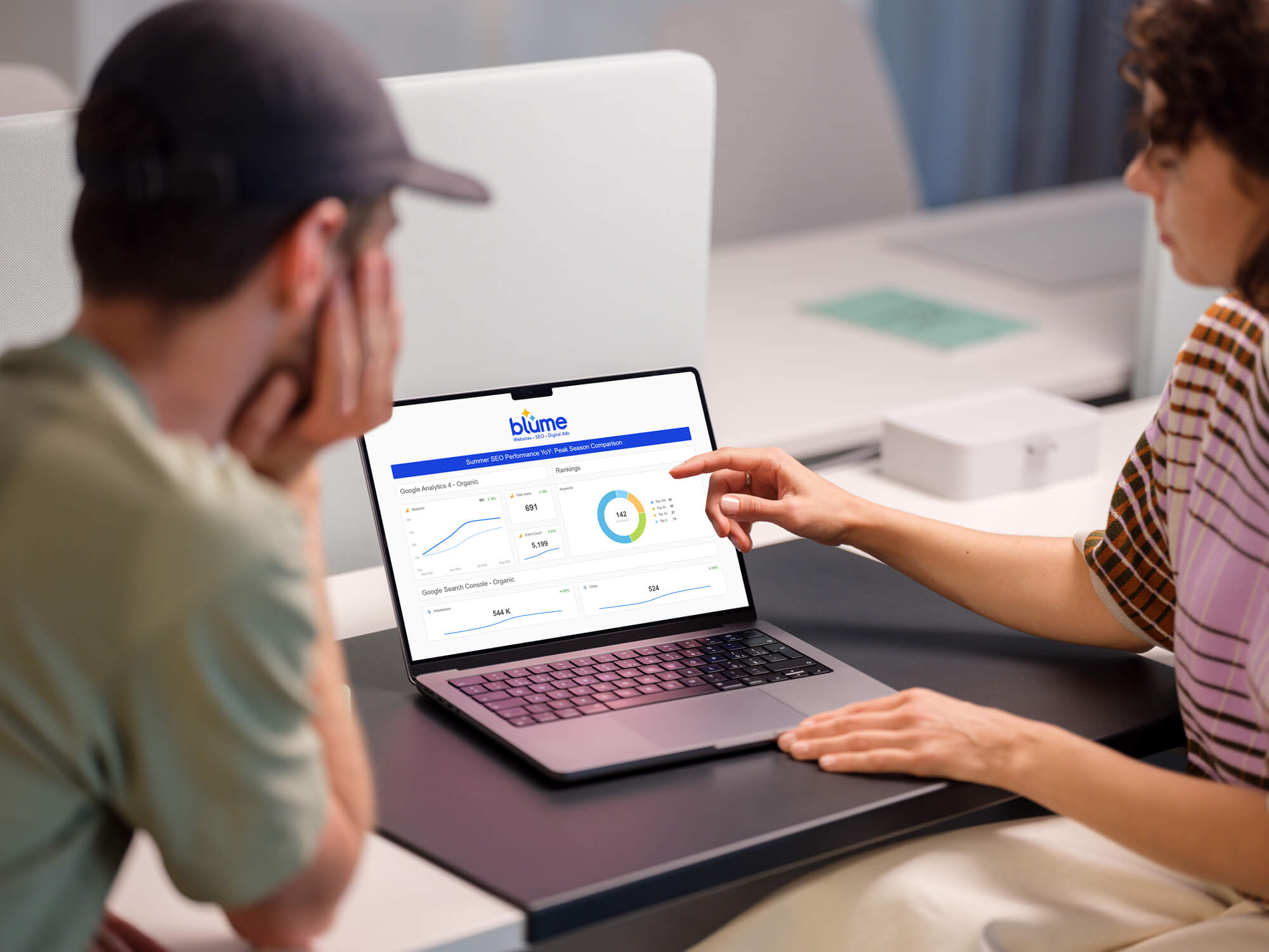 Two people sit at a desk looking at a laptop. One person points at the screen, which displays “blume,” a financial application dashboard with charts, graphs, and numerical data—ideal for an SEO company Detroit team optimizing performance insights.