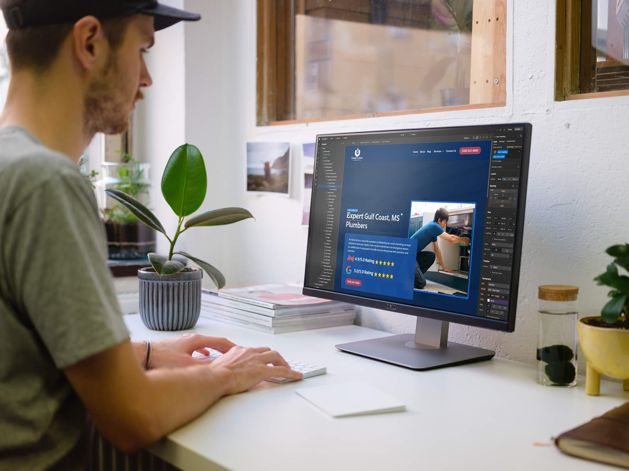 A person wearing a cap uses a keyboard and mouse at a desk with a monitor displaying a web design Alexandria LA interface. A potted plant, magazines, and a glass of water are arranged neatly on the desk.