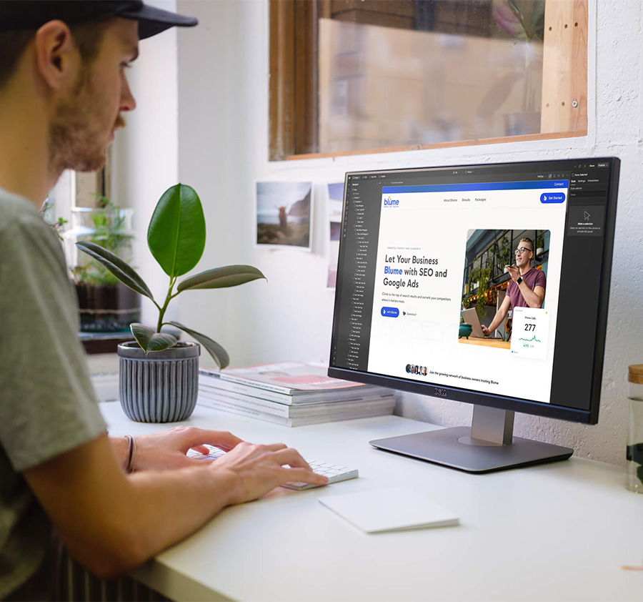 A person sits at a desk using a computer, viewing a website about SEO and Google Ads. There is a potted plant, stacked magazines, and an external keyboard on the desk near the monitor.