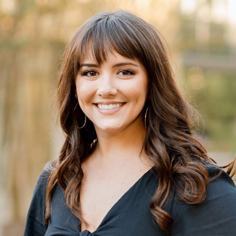 A woman with long brown wavy hair and bangs smiles at the camera. She is wearing a black top and hoop earrings, standing outdoors with a blurred background of trees.