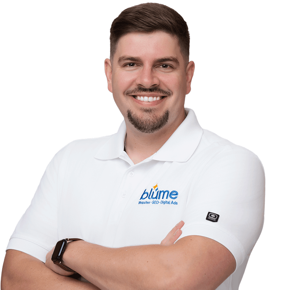 A person with short brown hair and a trimmed beard is smiling with arms crossed, wearing a white polo shirt with the "blume" logo and a smartwatch, standing against a plain white background.