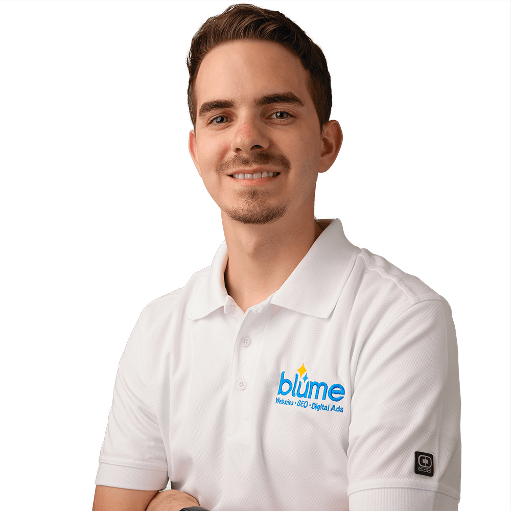 A man with short brown hair and a trimmed beard wears a white collared shirt with the "blüme" logo and stands with arms crossed, smiling at the camera against a plain white background.